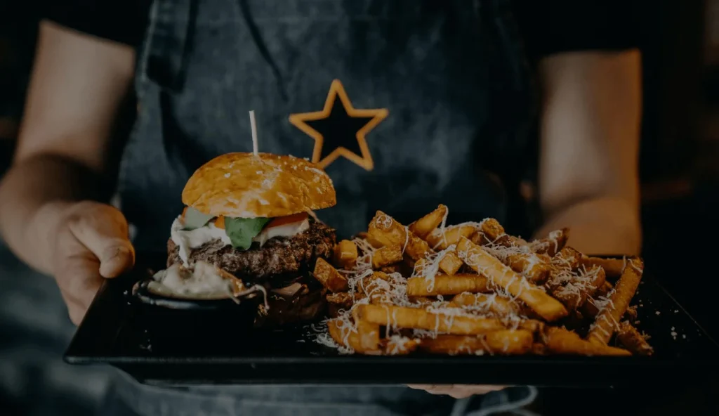 Tarjoilija kantaa tarjottimella burgeria ja ranskalaisia, esiliinassa tähtineula